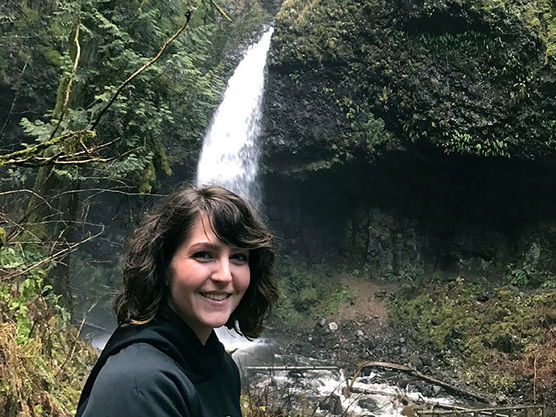 Person in a dark hoodie smiling in front of a waterfall and forested area.