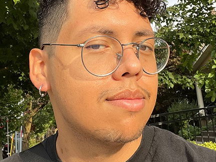 A person with curly hair and glasses outdoors in sunlight, wearing a black T-shirt.