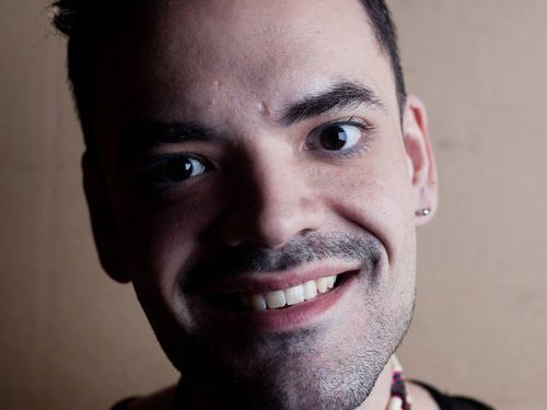 Close-up of a smiling person with short dark hair, earrings, and a beaded choker necklace against a beige background.