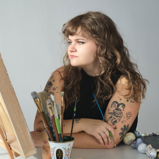 Young woman with wavy hair and tattoos sits at a table with art supplies, looking to the side.