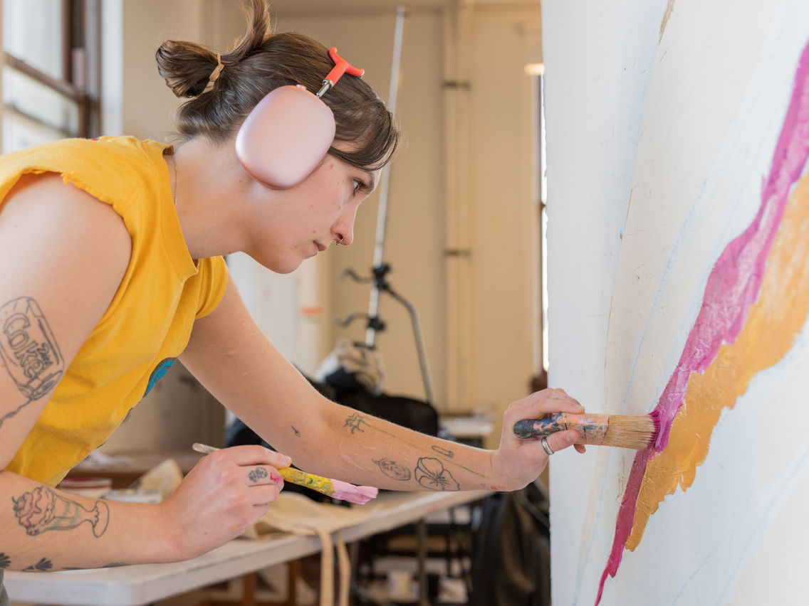 Person painting a mural with pink, orange, and light blue paint, wearing yellow shirt and pink headphones.