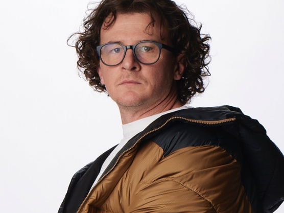 Person with curly hair and glasses wearing a brown puffer jacket in front of a white background.