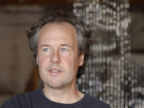 A man with salt-and-pepper hair and blue eyes stands indoors in front of a hanging metal rod sculpture.