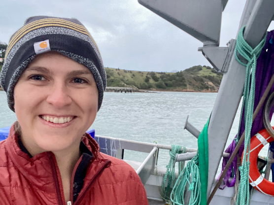 Person in a beanie and red jacket smiling on a boat with coastline in the background.