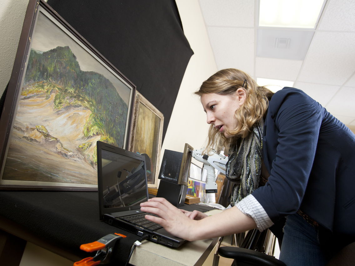 Woman leaning over a laptop with paintings and a microscope on the desk.