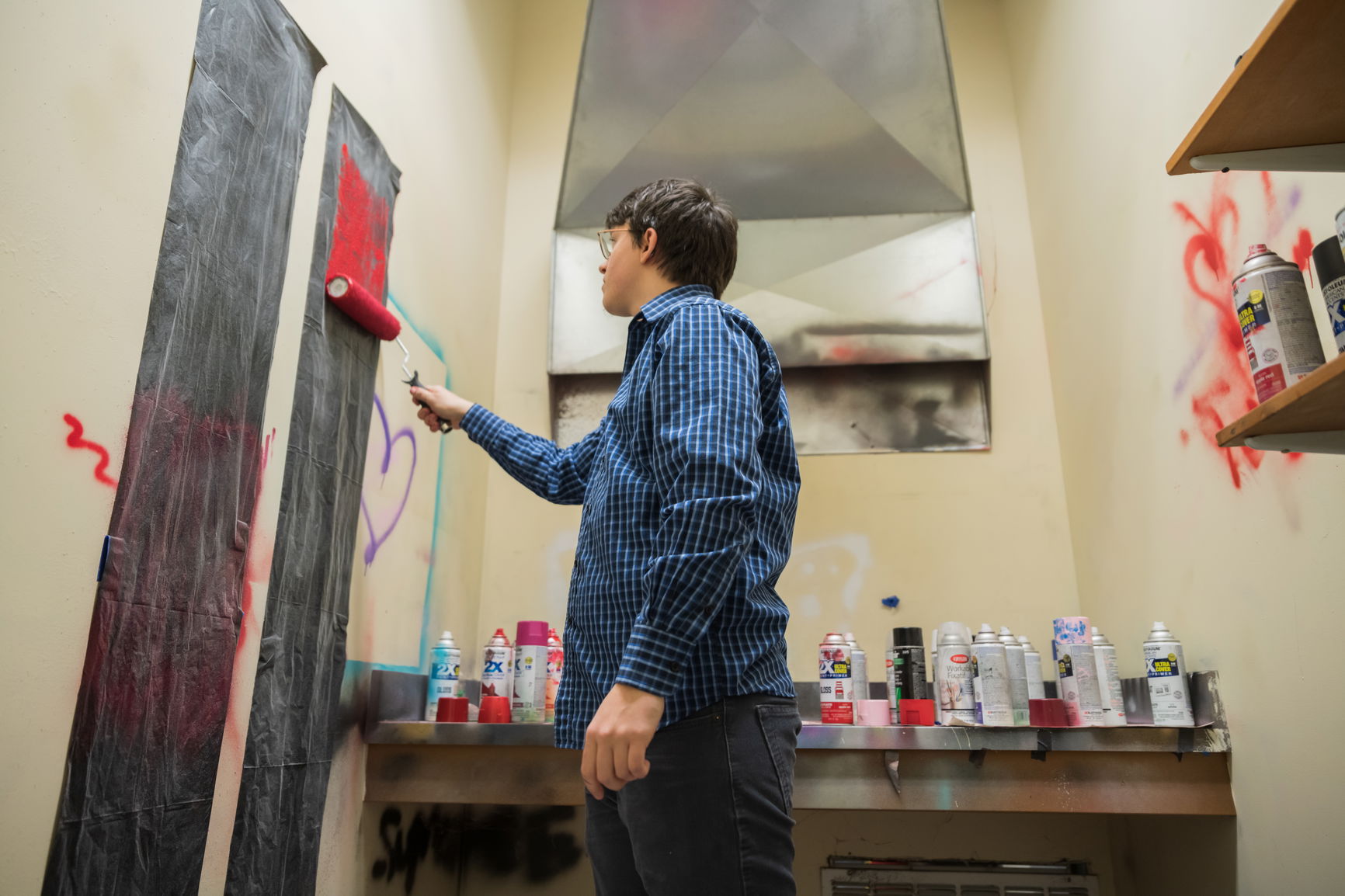 Person using a paint roller in a small spray painting booth with graffiti and numerous spray paint cans.