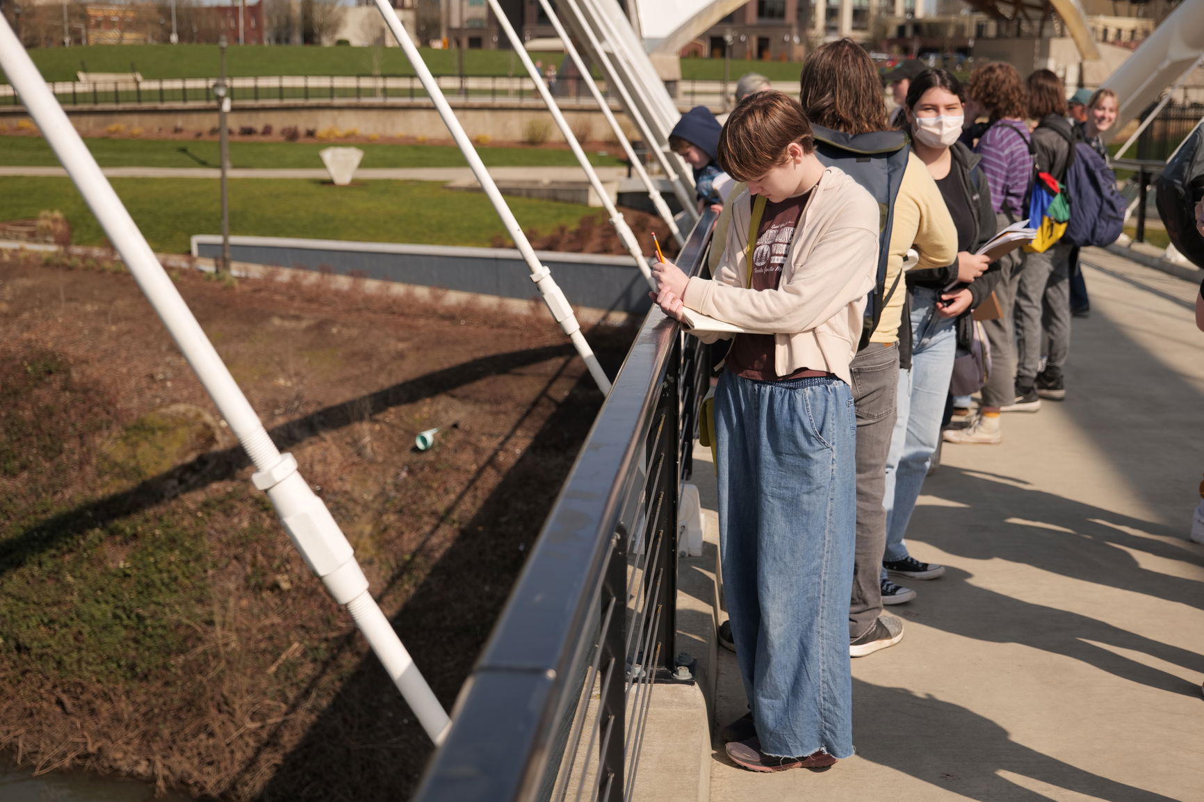 Group of people standing on a bridge, with one person in the foreground writing on a notepad.