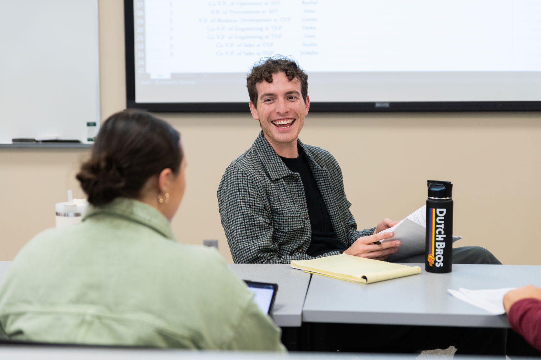 Young man smiling in a classroom, holding papers with a yellow notepad in front of him. Another person faces away from the ca