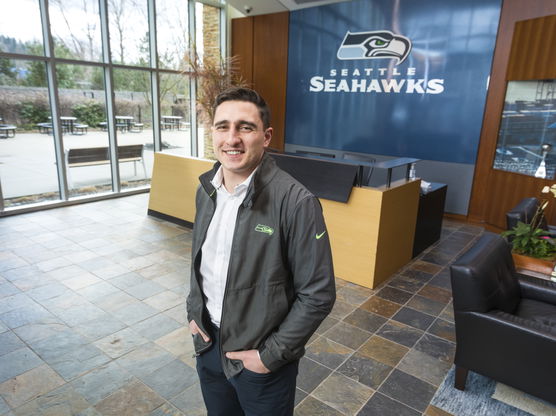 A man standing in a lobby with a Seattle Seahawks logo on the wall behind him.