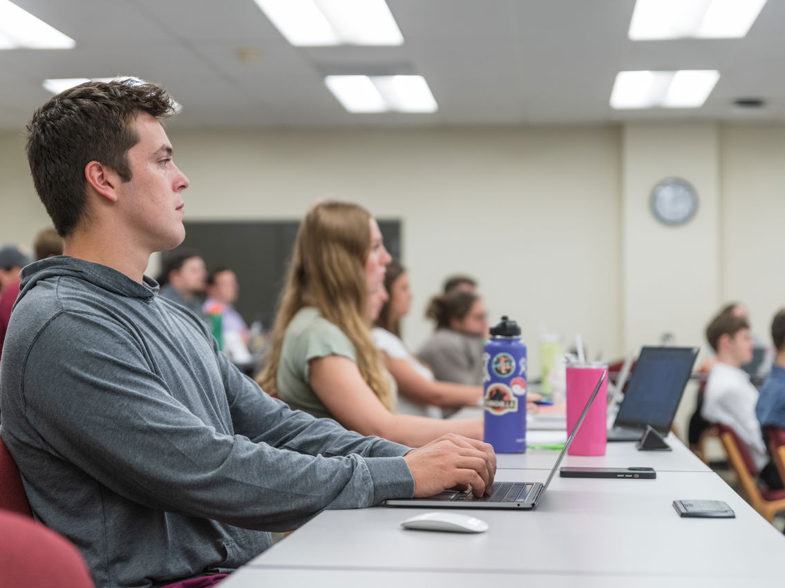 A man typing on his laptop in a classroom setting