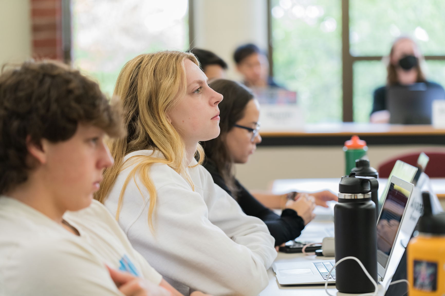 Students seated at desks with laptops in a classroom, appearing attentive.