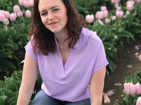 A young woman in a light purple blouse and blue jeans kneels among pink tulips in a field.