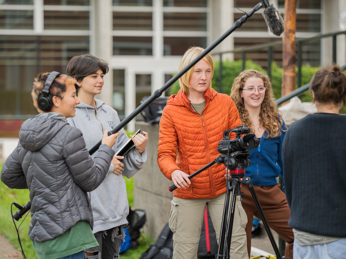 Five people engaged in an outdoor filming activity with a camera on a tripod, a boom microphone, and various equipment.