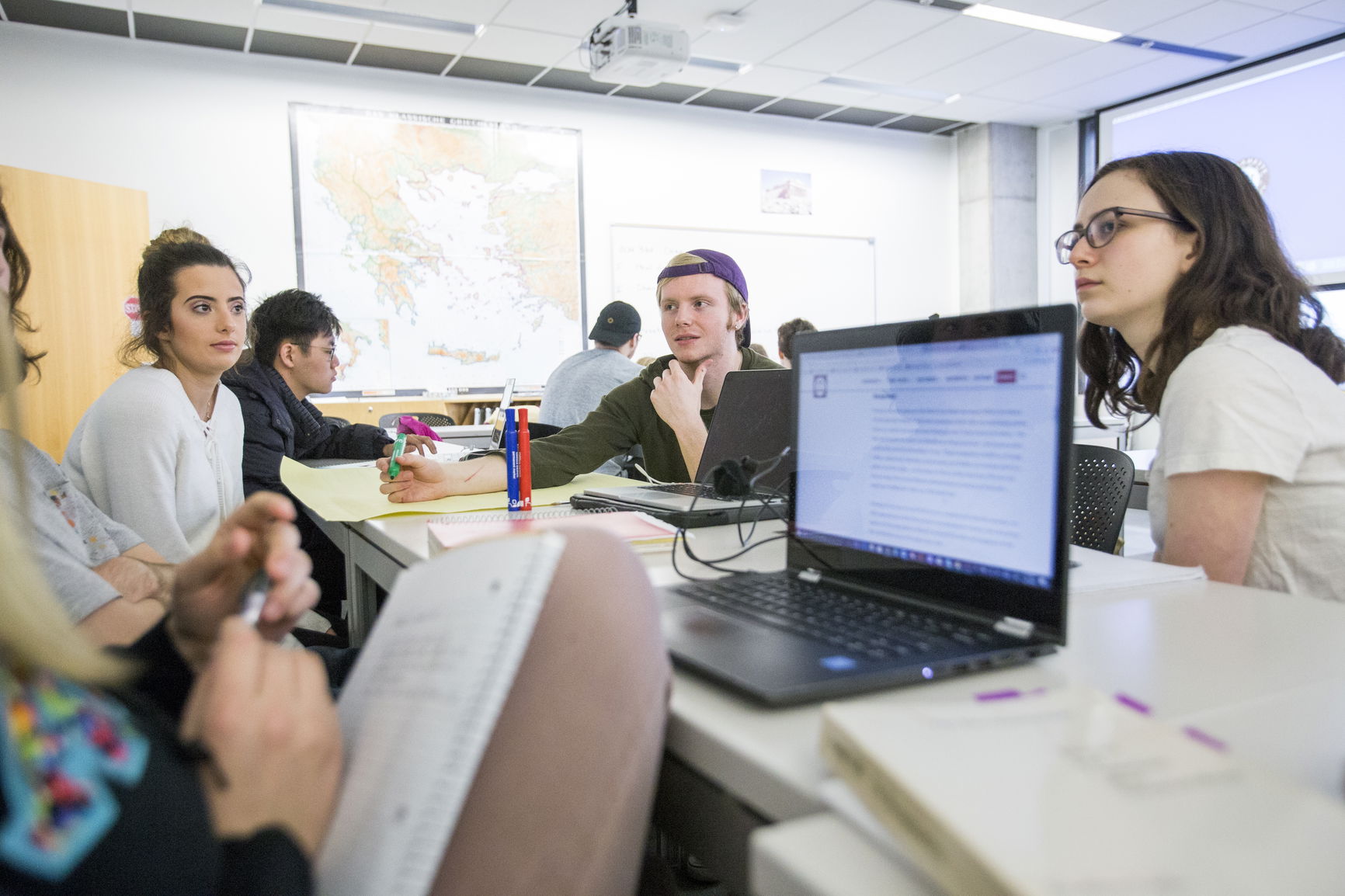 Students engage in a group discussion around a table with laptops, notebooks, and pens in a classroom with a map and whiteboa