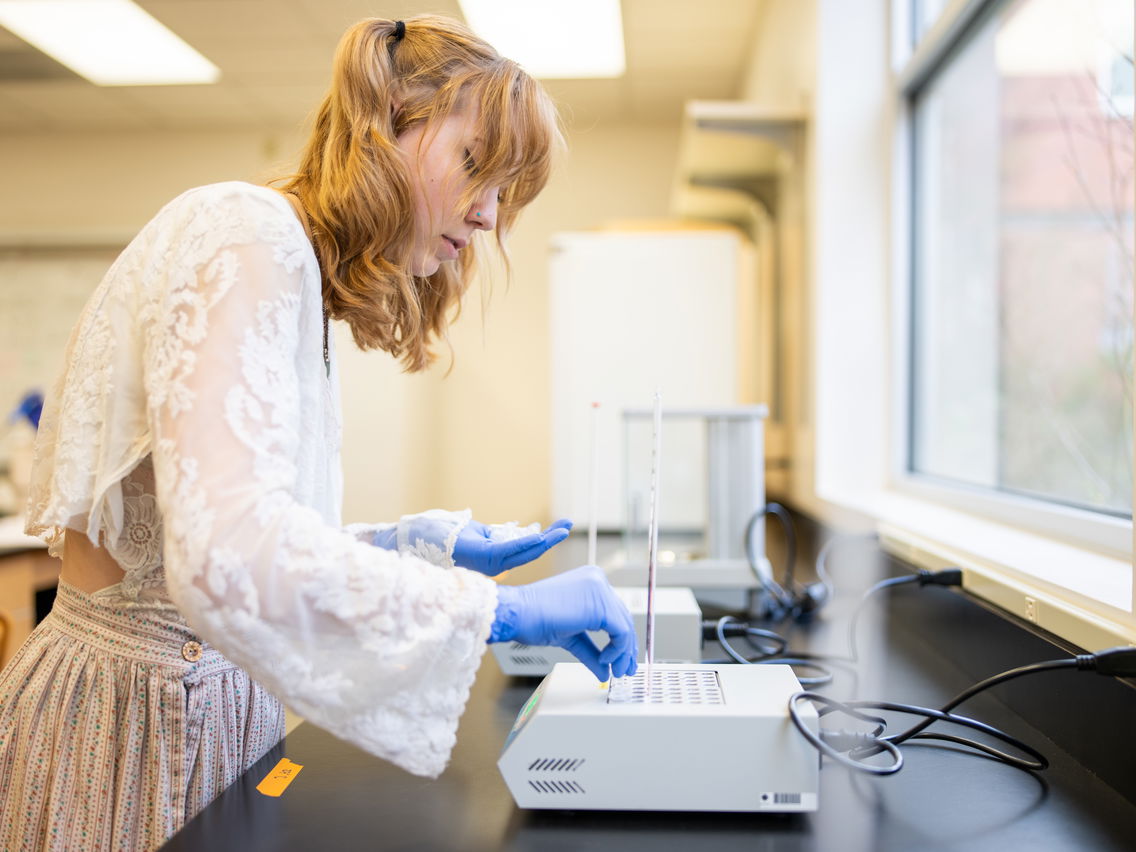 Young woman working in a lab with blue gloves, adjusting a gray lab machine.