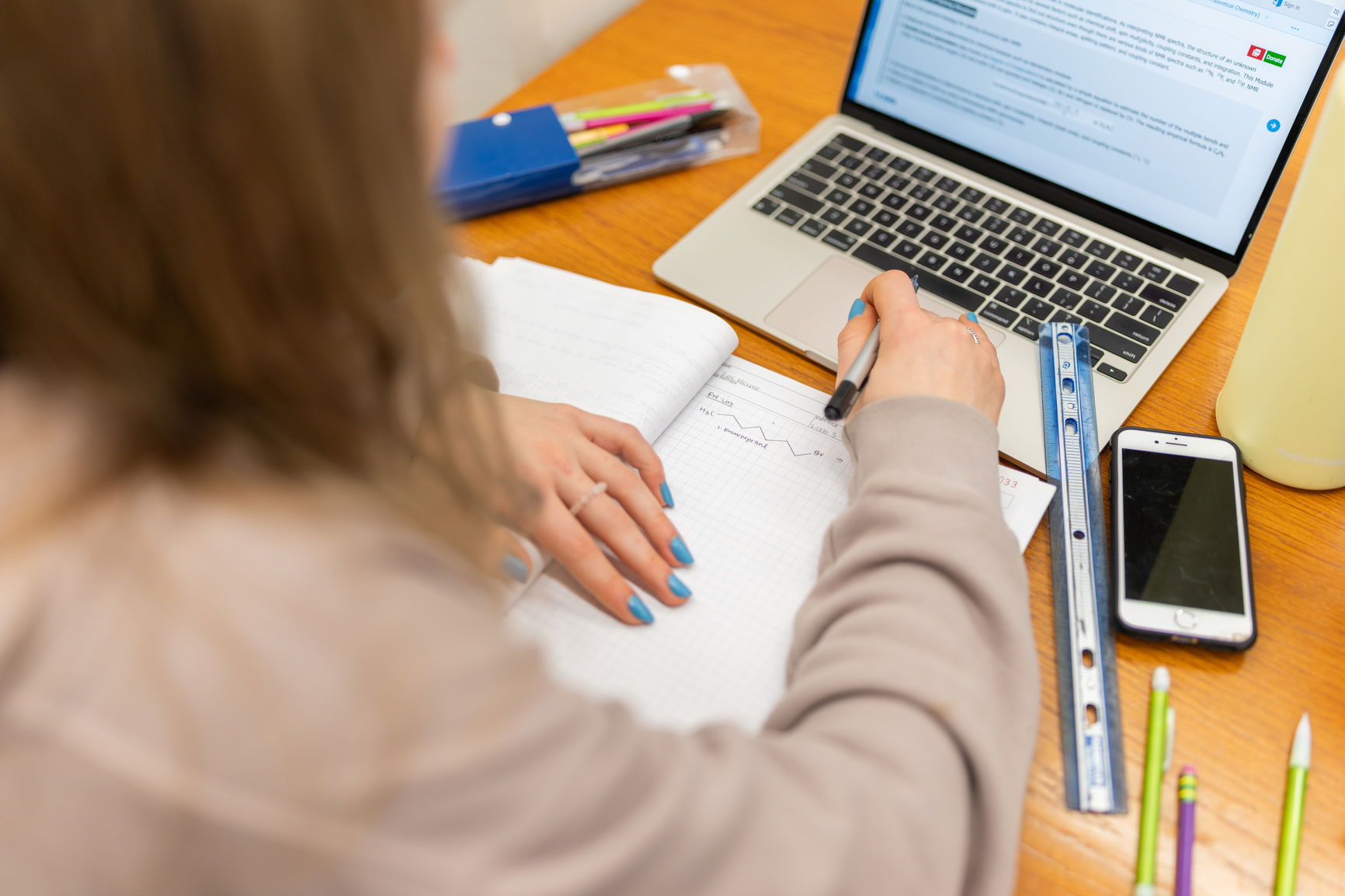 Person writing in a graph paper notebook at a desk with a laptop, rulers, smartphone, water bottle, and a pencil case.