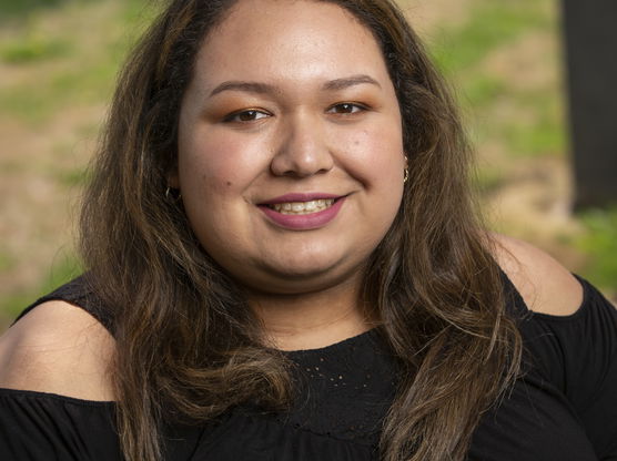 Close-up of a person with shoulder-length brown hair wearing a black shirt with cut-out shoulders, smiling slightly.