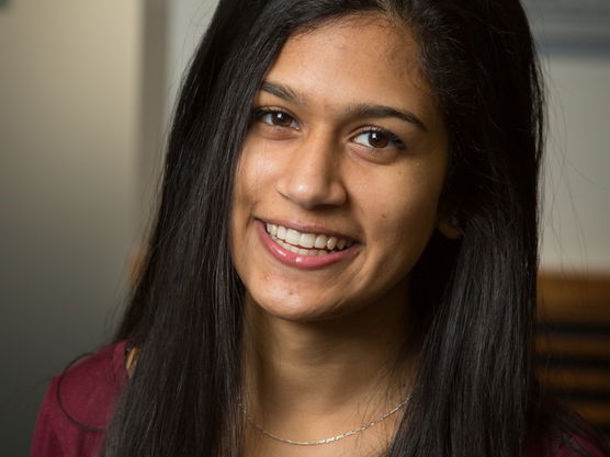 A young person with long black hair smiling, wearing a maroon top and a silver necklace. Blurred indoor background.