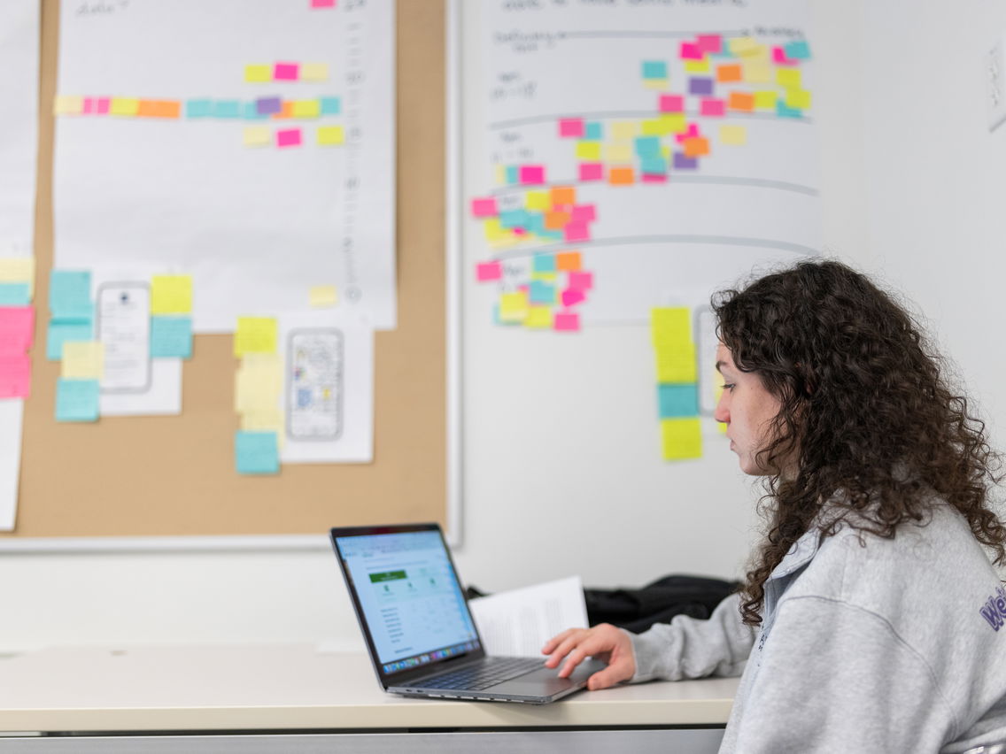 A young woman scrolling on her laptop in a classroom with plenty of sticky notes