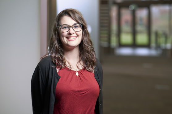 Young woman with glasses wearing a red blouse and black cardigan, smiling in an indoor hallway.