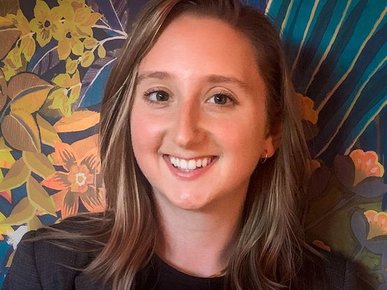 A smiling young woman with light brown hair in a blazer, against a colorful floral background.