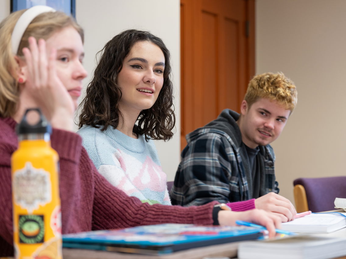 Three young adults in conversation, seated at a table with books and a yellow water bottle.