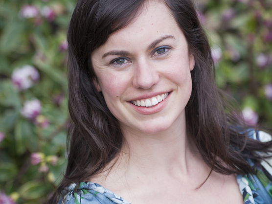 Smiling woman with dark hair in a blue patterned blouse, with a background of greenery and pink flowers.