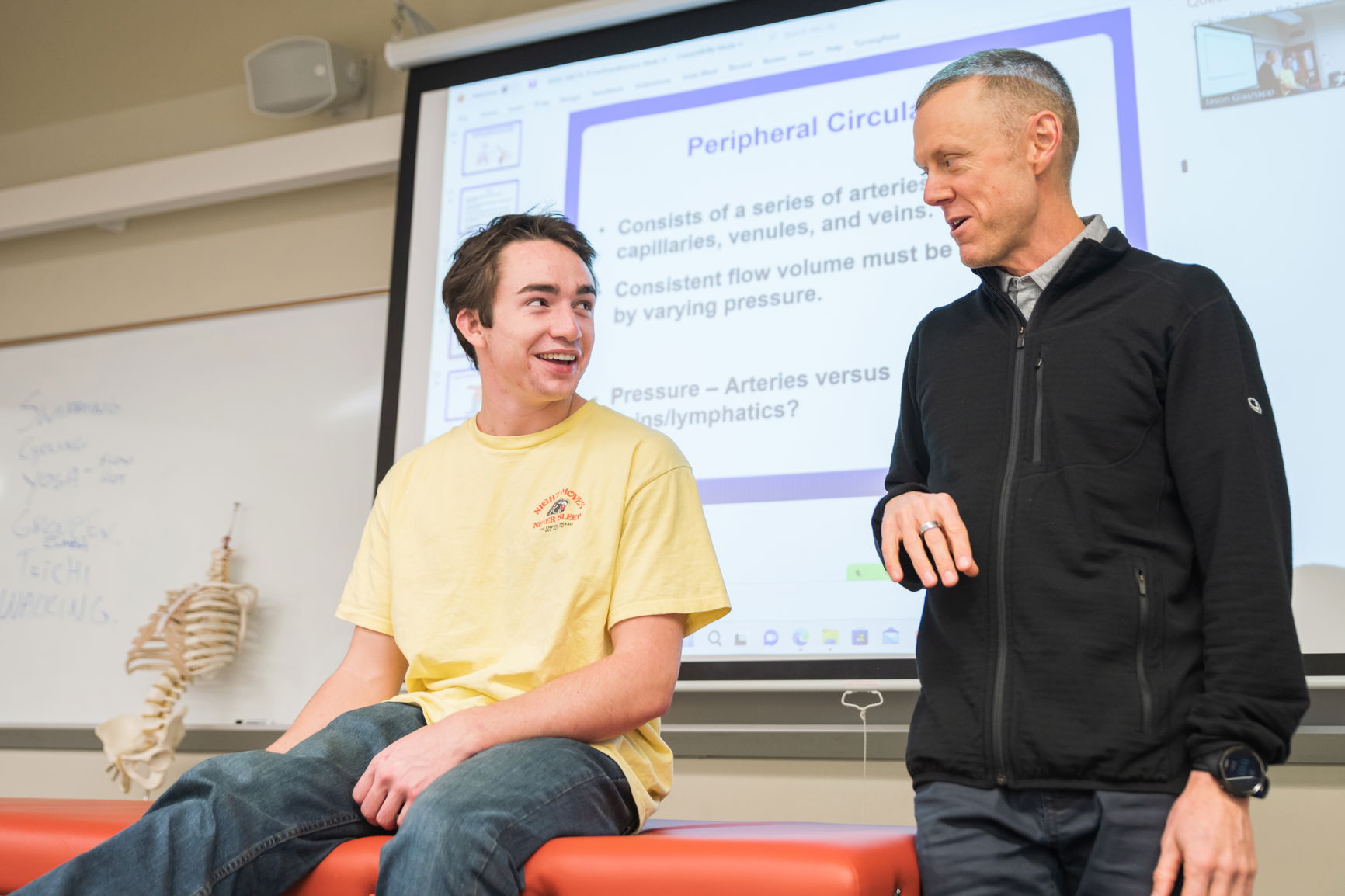 A professor next to a student who is on an exam table in the front of a classroom. 