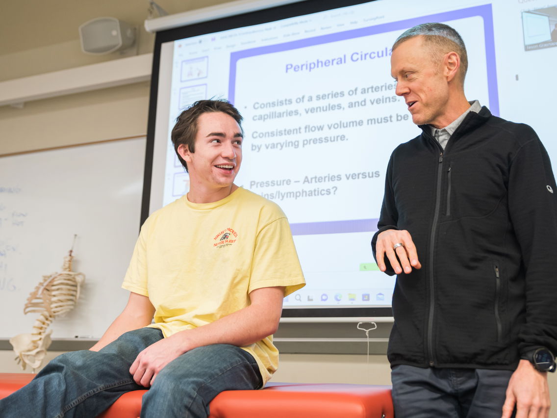 Professor standing next to a student sitting on an exam table in a classroom. 