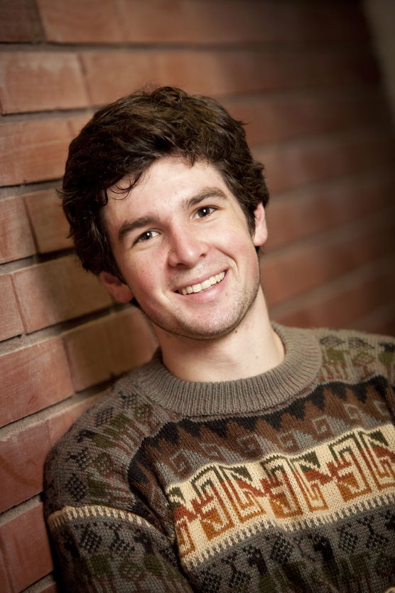 Young man leaning against a brick wall, wearing a patterned sweater, and smiling at the camera.