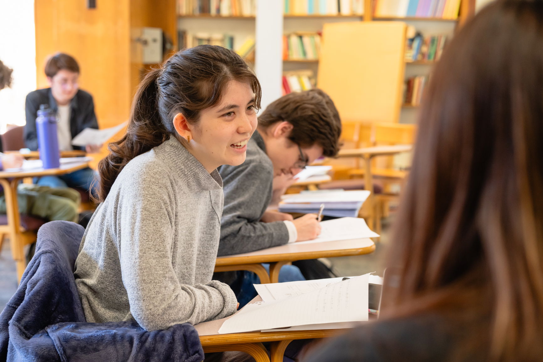 A brown haired young woman speaking to another person in a classroom.