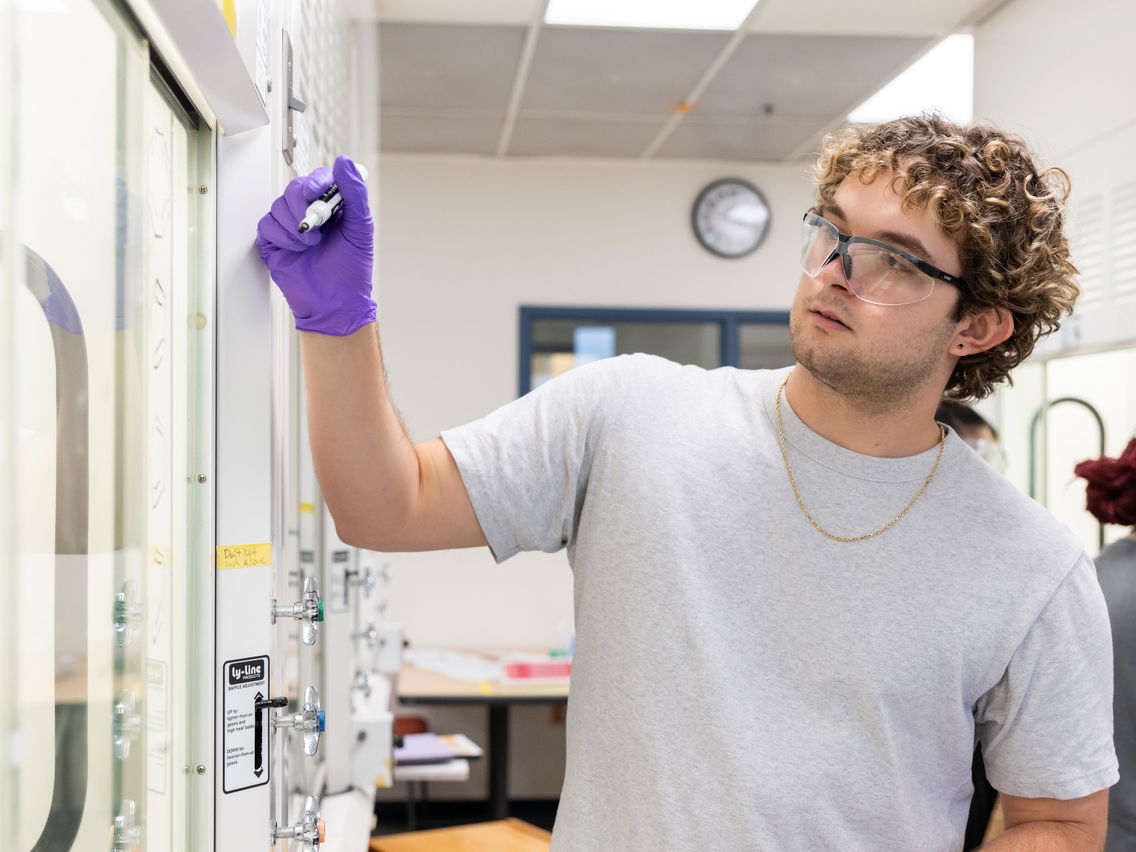 A student wearing goggles and writing on a white board in a chemistry lab. 