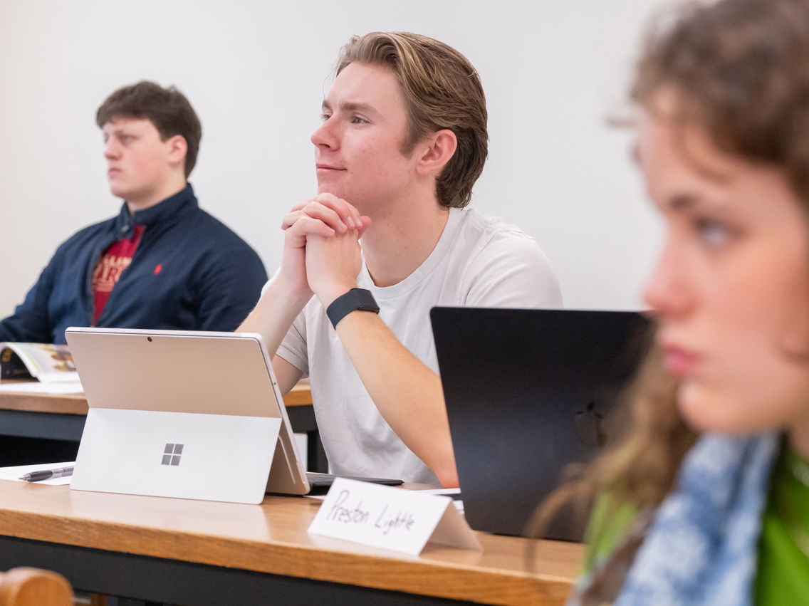 Two students sitting at a desk, listening to a lecture, with another student out of focus in the foreground. 