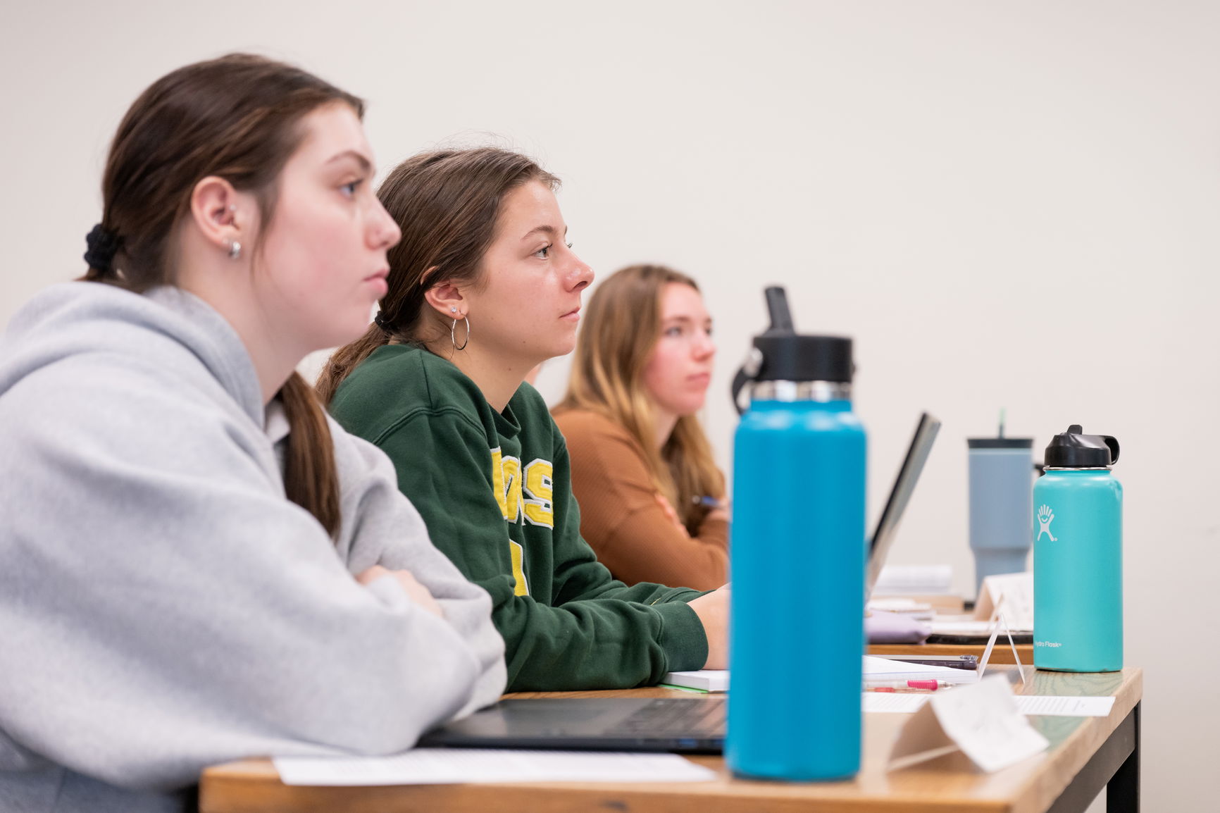 Three students sitting in a classroom with water bottles and laptops