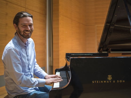 Person smiling while playing a black grand piano in a wooden-paneled room.