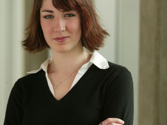 Young woman with short red hair, wearing a black sweater over a white collared shirt, standing with arms crossed.