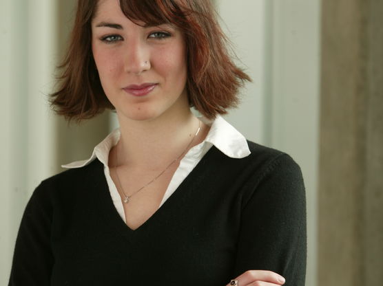 Young woman with short red hair, wearing a black sweater over a white collared shirt, standing with arms crossed.