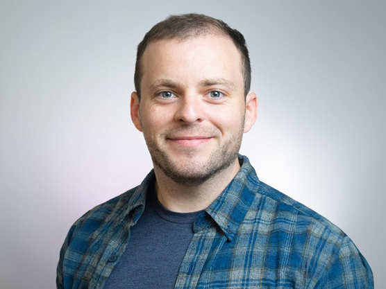 Man wearing a blue plaid shirt and a gray t-shirt, smiling at the camera.