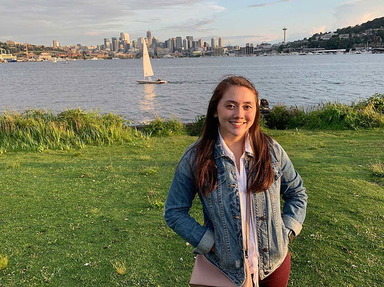 Woman in a denim jacket standing in front of a body of water with a sailboat and city skyline in the background.