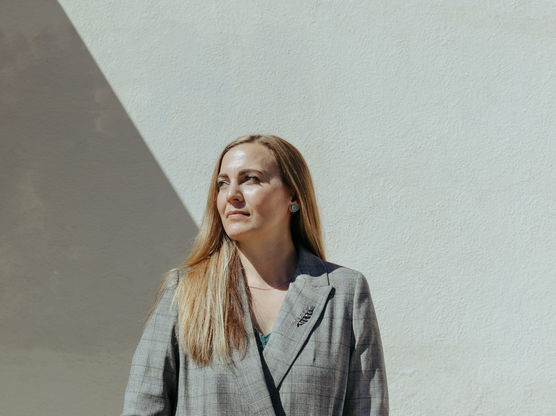 Woman in a gray blazer standing against a textured wall with a diagonal shadow.