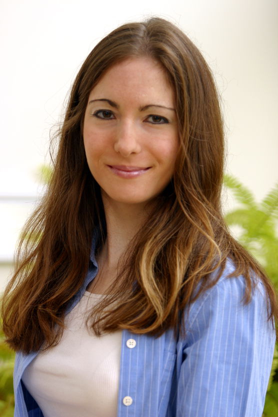 Portrait of a smiling woman with long brown hair wearing a light blue, striped shirt over a white top.