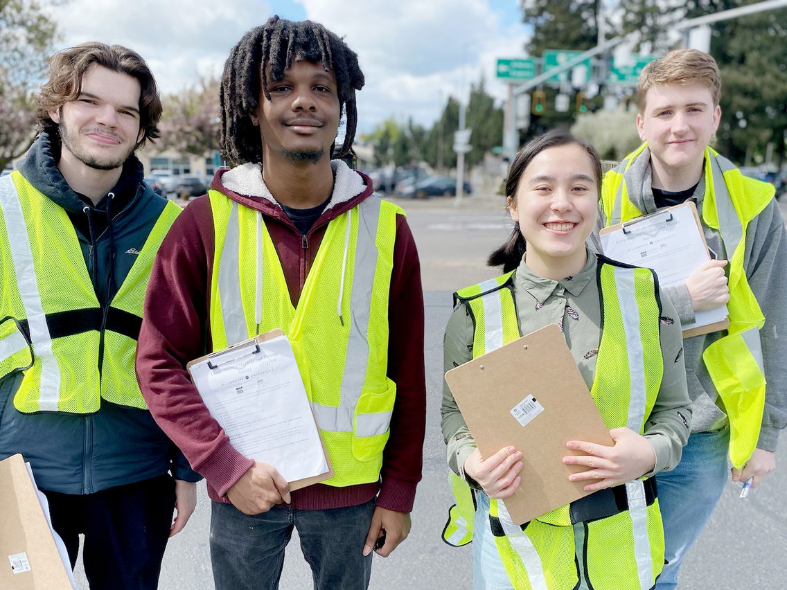 Four students standing outside wearing safety vests and carrying clipboards. 