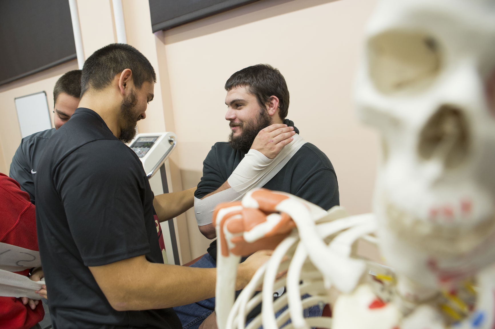 Man with arm in a sling being assisted by two others, with a skeleton model in the foreground.