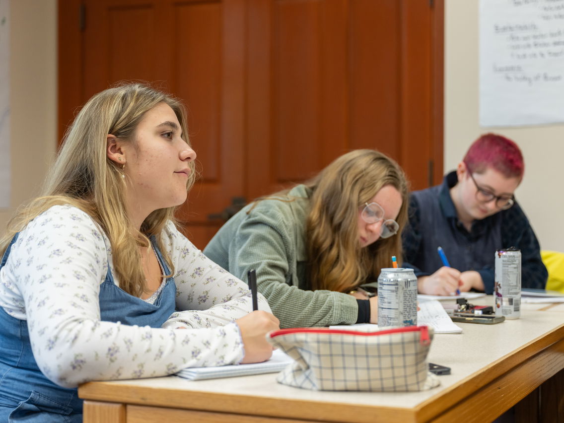 Three students sitting at a table listening to a professor. 