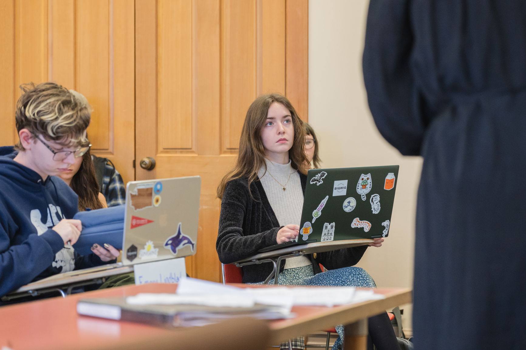 Students in a classroom with laptops, focusing on a young woman looking forward and a young man reviewing a folder.