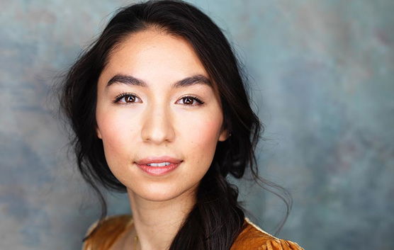 Portrait of a young woman with long, dark hair, looking directly at the camera against a bluish-gray background.